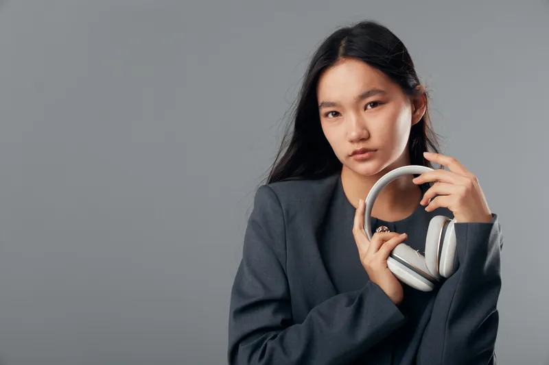 Young woman in gray blazer holding white wireless headphones with serious expression on gray background. Portrait of confident female, technology accessory and casual style fashion.