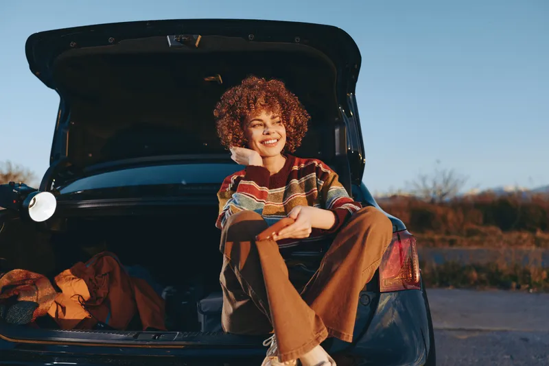woman car trunk mother child outdoors lifestyle road travel, smiling curly hair, mother hugs child in open trunk, warm sunset light, casual adventure, packed luggage in vehicle on rural road