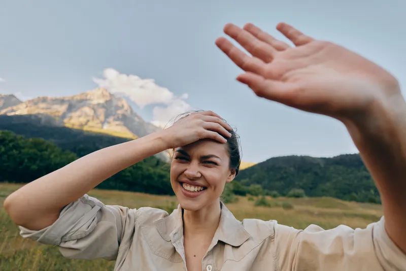 Young woman with short hair smiles in a sunlit field with distant mountains behind her as she shields her eyes with one hand, enjoying a carefree moment outdoors.