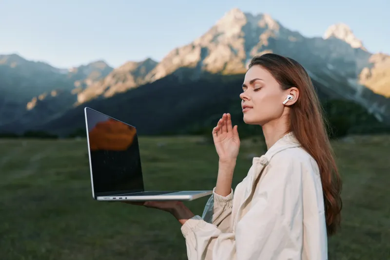 An outdoor professional woman holds a laptop in one hand, wearing wireless earbuds, gazing thoughtfully ahead in a vast scenic mountain landscape, conveying focus and calm.