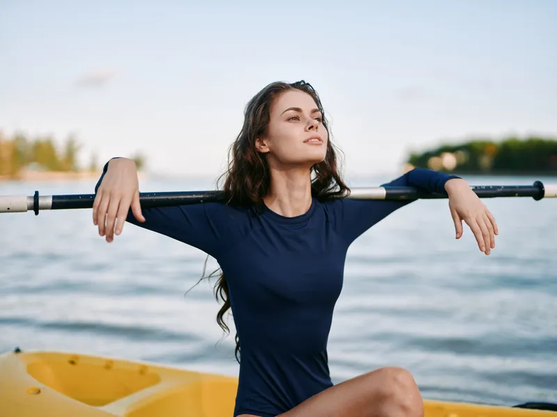 A serene portrait of a woman resting with an oar across her shoulders on a calm lake, wearing a navy swimsuit and basking in gentle sunlight.