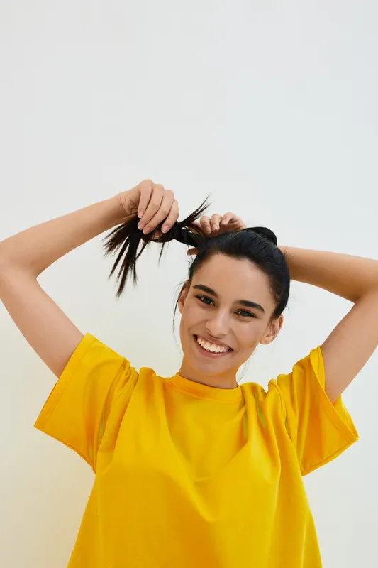 Smiling woman in yellow shirt tying hair back, cheerful young adult with dark hair and bright outfit, isolated on light background, natural expression and casual style.