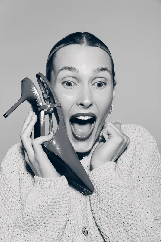 Excited white woman with fair skin holding a high heel shoe near her face, wearing knitted sweater, studio portrait.