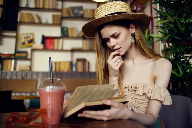 Симпатичная женщина в шляпе с книгой в руках читает, отдыхая в кафе во время отпуска