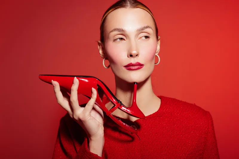 Woman in red sweater holds red high heel shoe near face on red background. Fashion concept with matching colors, style, and confident female model posing in studio lighting.
