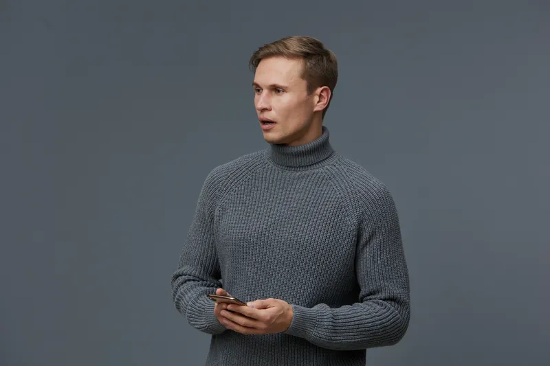 Man wearing gray turtleneck sweater holding smartphone, looking sideways, isolated on plain gray background. Studio portrait of young adult male with serious expression, casual clothing, focused on te