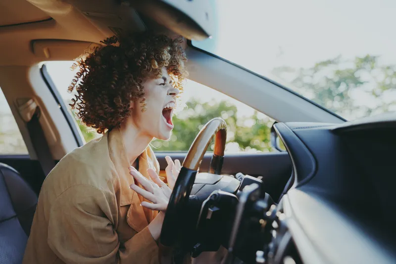 Woman driving in a car, casual lifestyle moment, she screams with wide eyes, retro film color casting, candid indoor scene, daytime light on dashboard, bold emotional gesture