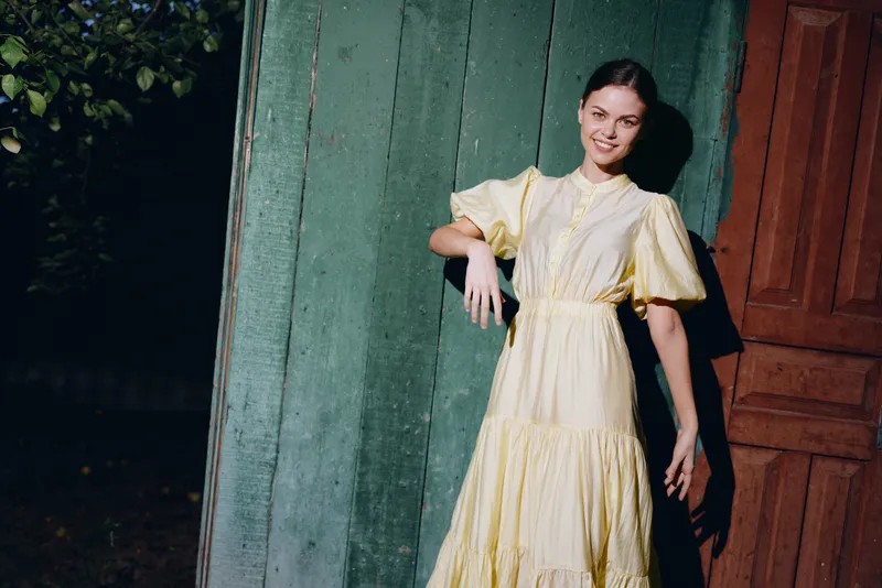 Woman wearing a yellow dress poses beside a teal wall and wooden doorway, creating a vintage fashion portrait with soft lighting and a calm outdoor mood.