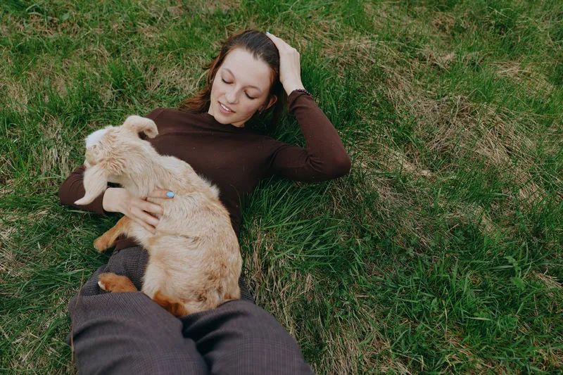 Relaxed woman lies on green grass outdoors, cuddling a young goat kid, sharing a gentle moment of companionship and calm, natural light, casual lifestyle scene with soft shadows, breeze, rural