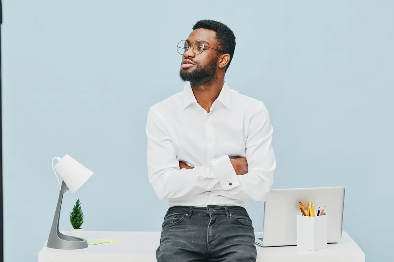 Confident young man with glasses, black hair, and casual white shirt standing with crossed arms behind a desk in a modern office with a minimalist light blue background, creating a professional workpl