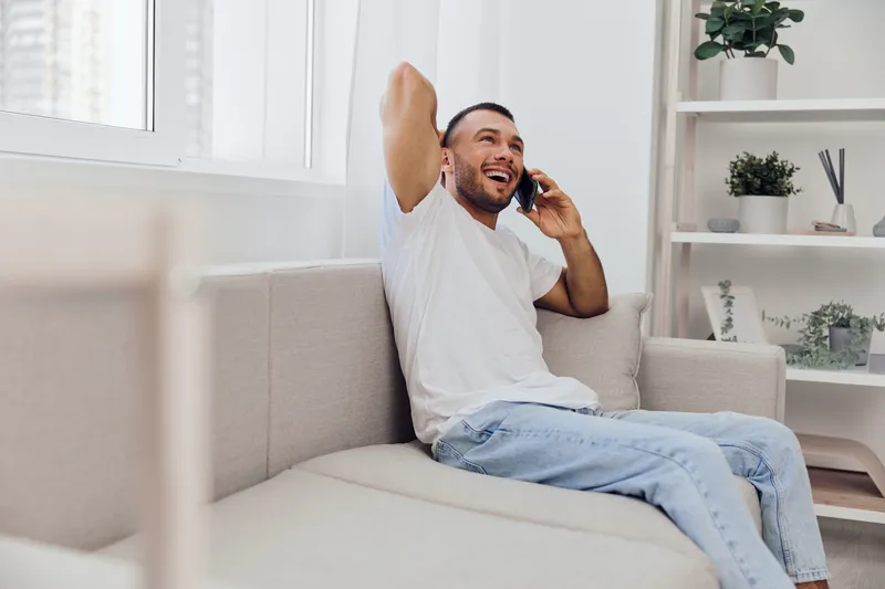 Smiling man enjoying a phone call, relaxed in comfortable home environment, expressing joy and connection. Perfect representation of soft masculinity and mental wellness.