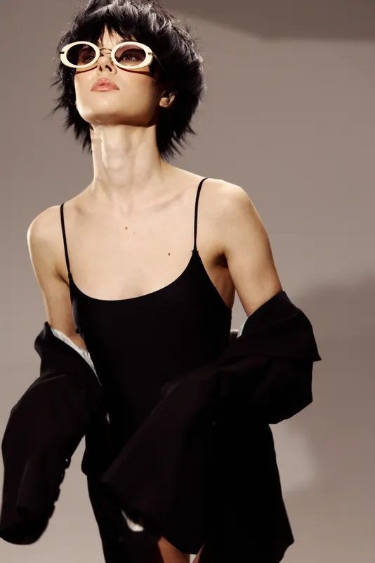 Stylish woman wearing sunglasses with short hair and black outfit posing confidently indoors with a neutral background