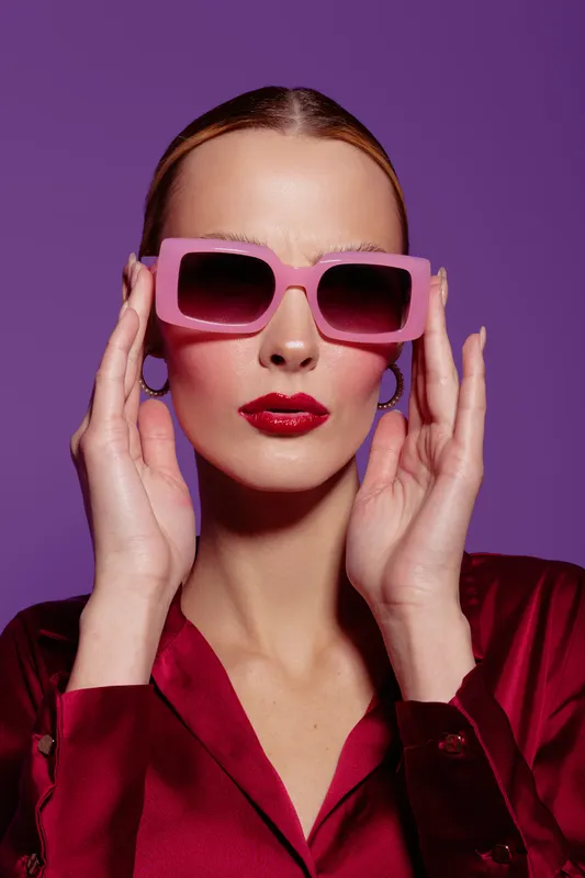 Stylish woman with pink sunglasses and red lipstick poses against purple background. Fashion portrait with trendy accessories and confident expression in studio lighting.