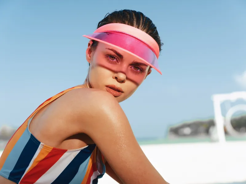 Vibrant portrait of a sun kissed woman at the beach, wearing a striped swimsuit and pink visor, glistening skin, relaxed pose, dynamic light, and a lively summer mood.