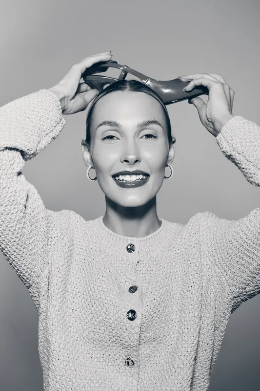 Young white woman with fair skin smiles while holding high heel shoes on her head. Casual cozy sweater, hoop earrings and joyful expression in black and white photo.