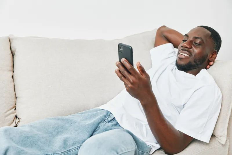 Smiling young Black man lounging on a sofa, enjoying his smartphone while wearing casual white t shirt and blue jeans, relaxed atmosphere in a cozy home setting