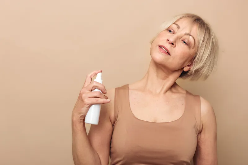 Mature woman with short blonde hair sprays facial mist on her neck, wearing beige top against a matching background. Concept of skincare, hydration, self-care, and beauty routine.