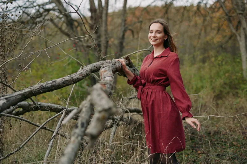 Authentic portrait of a woman in a burgundy dress outdoors, leaning on a weathered branch, conveying calm, warmth, and genuine connection with nature.