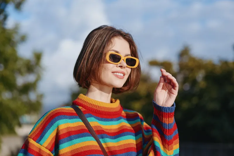Woman in her mid 20s smiling in a colorful striped sweater and yellow sunglasses, short bob haircut, casual outfit outdoors in a sunny urban park, looking up and enjoying travel lifestyle.