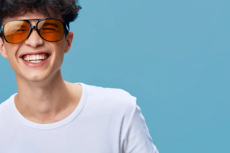 smiling young man wearing orange sunglasses and a white shirt, cheerful expression, curly hair, studio shot with blue background, casual style, summer vibe