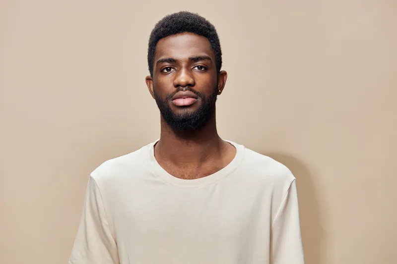 Portrait of a young Black man wearing a casual beige t shirt with a neutral expression against a minimalistic brown background, showcasing modern urban style and natural beauty