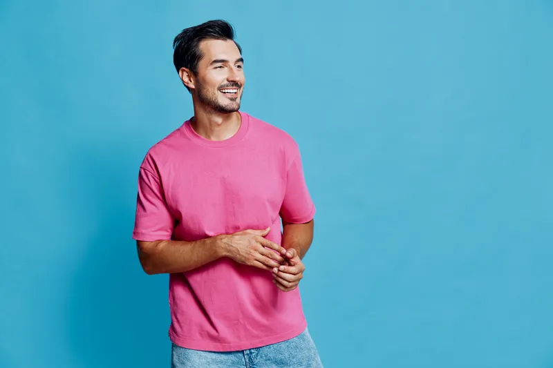 Happy cheerful young man wearing a pink t-shirt, standing against a solid blue background, smiling and enjoying the moment, portraying a lively lifestyle concept.