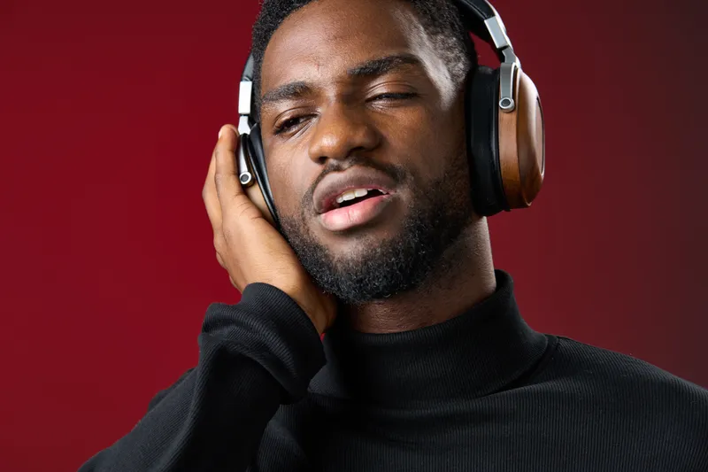 Young Black man enjoying music with headphones on, expressing emotions against a deep red background, showcasing modern style and a relaxed demeanor