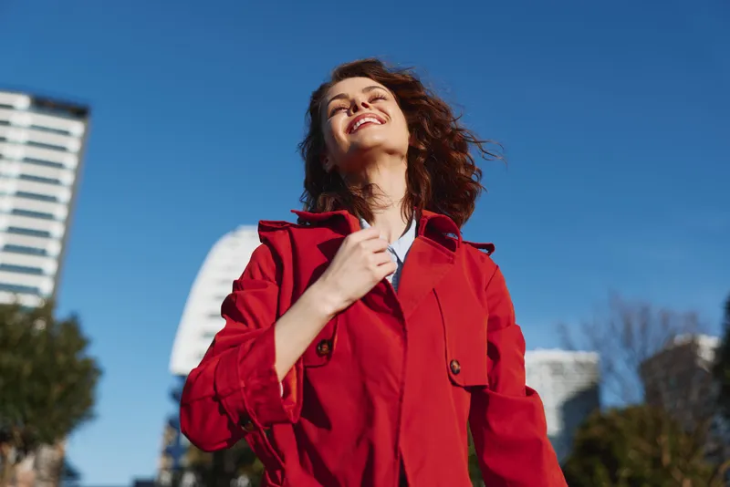 Smiling woman in a red coat outdoors, looking up with a carefree expression. Bright blue sky and urban background convey confidence, fashion, and wellness.