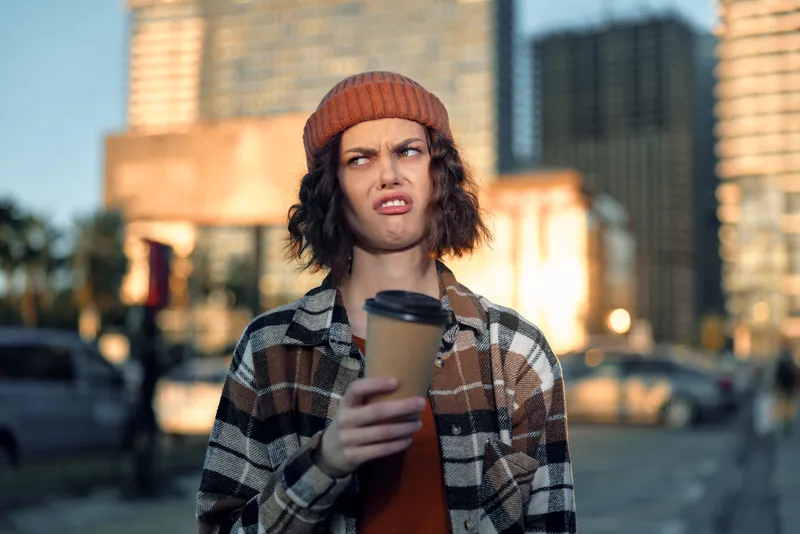 Person coffee expression city beanie plaid portrait urban authenticity candid lifestyle emotional storytelling golden hour glow young adult grimace holding takeaway cup on sidewalk in moody street