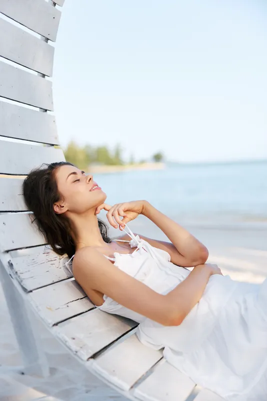 Relaxed woman lounges on a curved white wooden bench by a sunlit beach, eyes closed, enjoying a gentle sea breeze and peaceful mood in a seaside retreat.