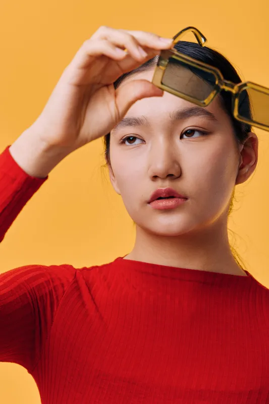 Young woman with smooth skin holding oversized sunglasses above her face isolated on yellow background. Portrait of thoughtful female model wearing red top with natural makeup and expression.