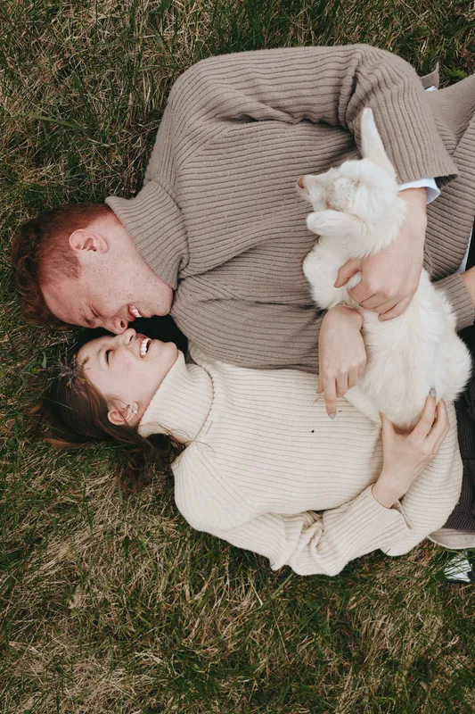 Candid couple lying on grass, cozy sweaters, smiling and cuddling a curious goat kid outdoors, warm sunlight, relaxed lifestyle moment, natural colors, rustic background, tender connection, gentle