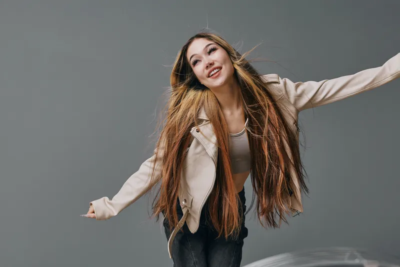 Young woman with long hair wearing beige jacket and gray top posing happily with arms outstretched against gray background in studio. Smiling, casual style and confident expression.