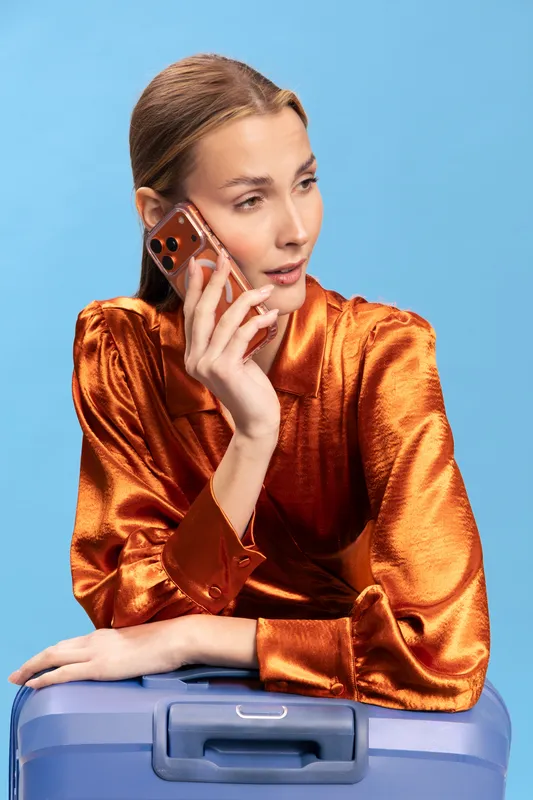Young woman in shiny orange blouse holding smartphone to ear leans on blue suitcase against blue background. Concept of travel, communication, and modern lifestyle with casual elegance.