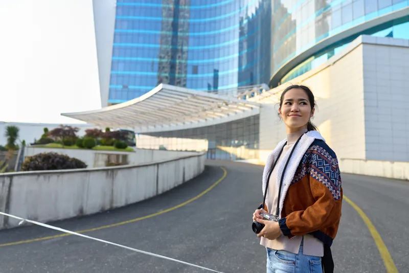 Woman with camera standing outdoors near modern architecture, urban walkway, casual jacket, and cheerful expression, ready for travel and lifestyle photography use.