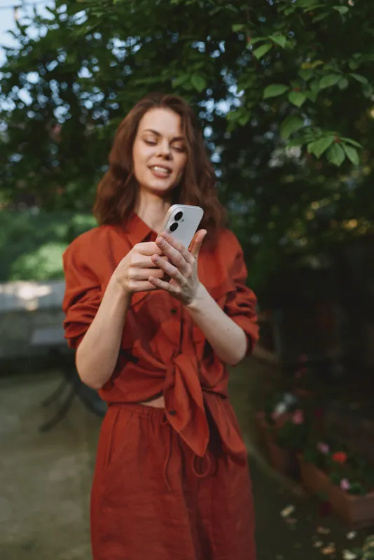 Casual lifestyle portrait of a joyful woman in an orange dress outdoors, checking her smartphone amid green greenery, relaxed smile and natural candid mood