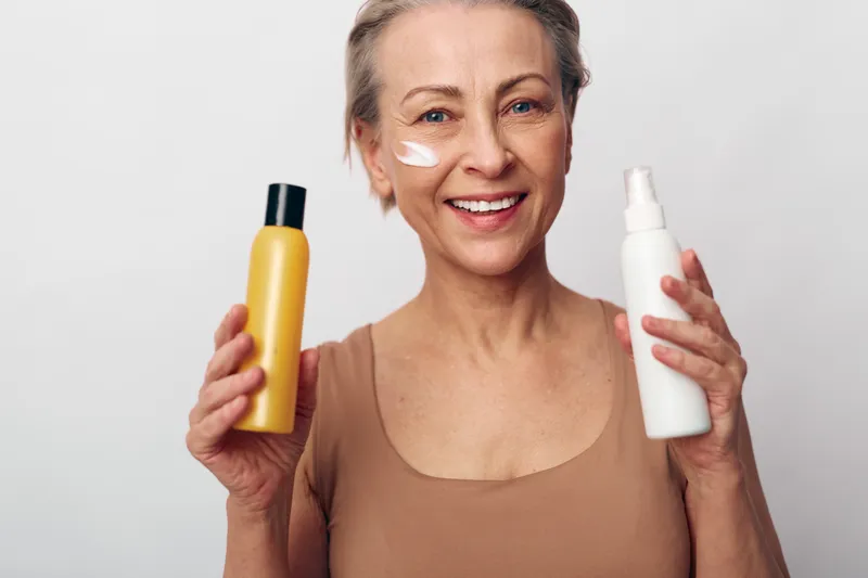 Smiling mature woman with short hair holds two cosmetic bottles and applies cream on cheek, promoting skincare, beauty routine, and healthy lifestyle on neutral background.