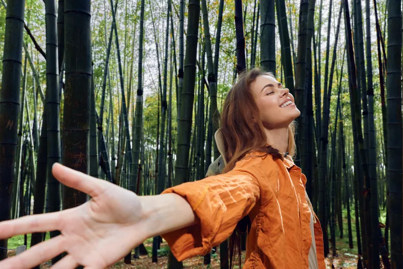 Woman in bamboo forest smiling with outstretched arms, enjoying nature and freedom. Portrait of relaxed young adult in orange jacket, peaceful outdoor moment and joyful breath.