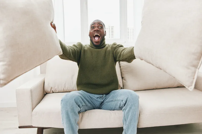 Excited man with pillows showing joy in a cozy living room, wearing a green sweater and jeans, bright atmosphere highlighting comfort and happiness