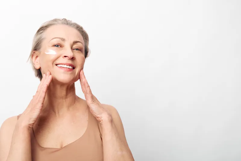 Smiling mature woman applies facial cream, showing skincare routine with natural makeup on clear skin. Concept of aging beauty, wellness, self-care, and healthy lifestyle in bright studio.