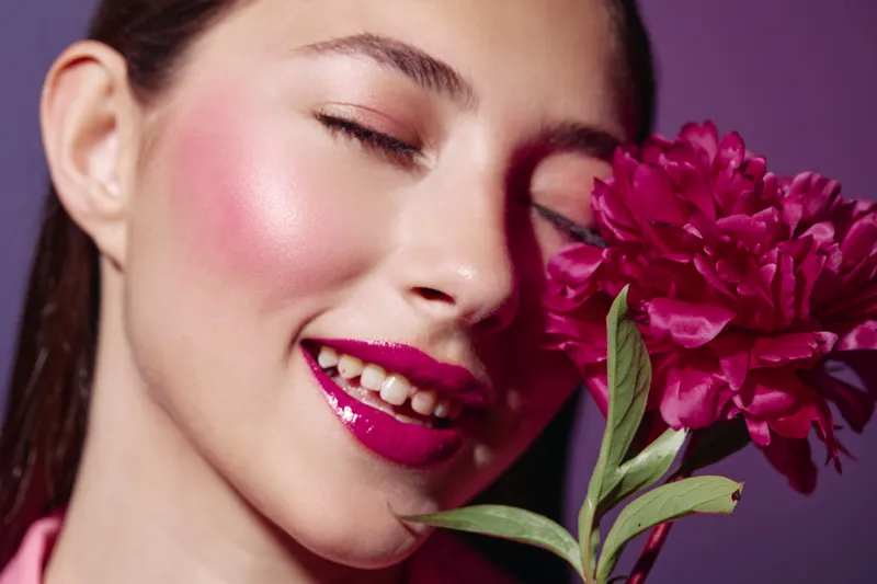 Close-up portrait of a young woman with flawless glossy makeup and bright magenta lips smelling a vivid pink flower with closed eyes. Concept for beauty fashion and creative aesthetics.