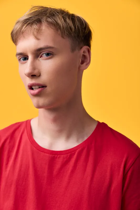 Young man in a red shirt against a warm yellow background, close up, neutral expression with a subtle smile, studio fashion shot, bold color contrast and clean composition