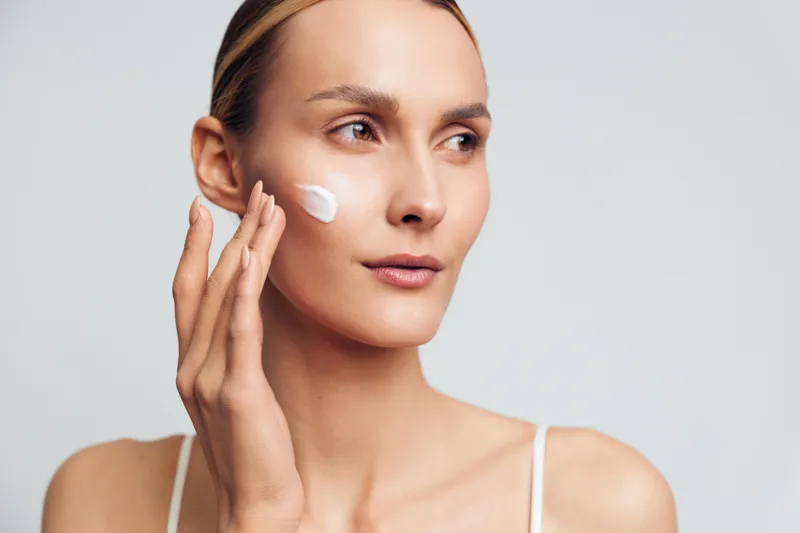 Young woman applies moisturizing cream on her cheek with fingers, shows natural skin care routine, healthy complexion and gentle facial treatment on white background.