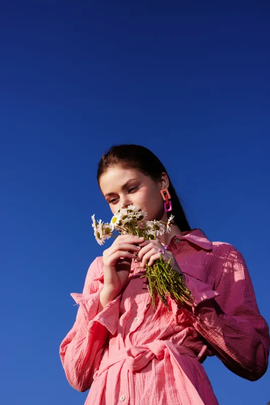 Young woman in pink dress smelling wildflowers against a clear blue sky, radiating joy and serenity, perfect for seasonal or nature themed projects