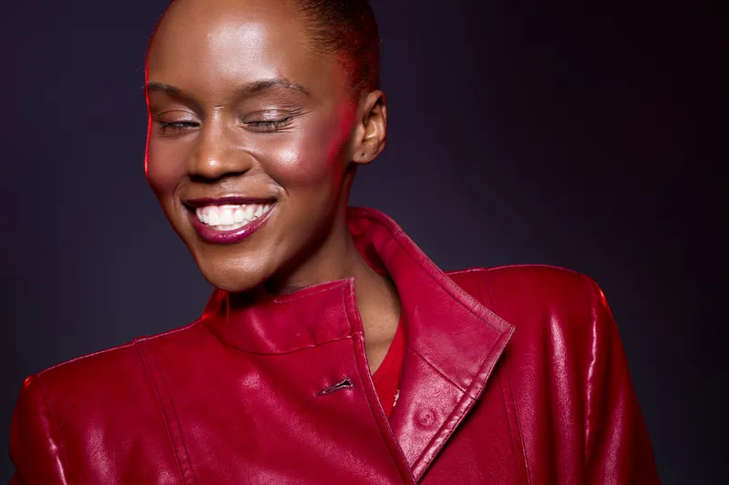Smiling black woman with dark skin wears shiny red leather jacket, eyes closed, showing joyful expression against dark background. Concept of fashion, beauty, and positive mood.