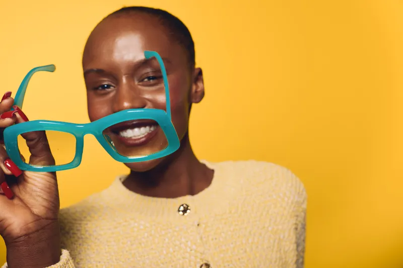 Smiling black African woman with dark skin holds large blue glasses in front of face against bright yellow background. Concept of beauty, fashion, and joyful lifestyle with vibrant colors.