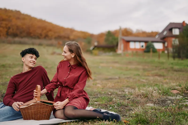 A young couple enjoys a relaxed countryside picnic on a blanket with a wicker basket, sharing a genuine moment that conveys authenticity and credibility in a natural everyday connection.