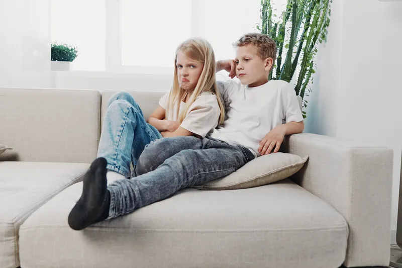 Upset girl and concerned boy sitting apart on beige sofa in bright living room. Emotional conflict and childhood tension concept with natural light and casual clothing.