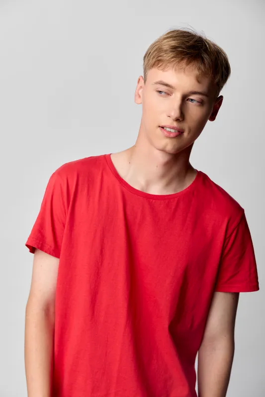 Young man wearing a red t shirt stands in a studio, relaxed pose, soft lighting, casual fashion, everyday style, contemporary look without branding, natural expression, clean background.