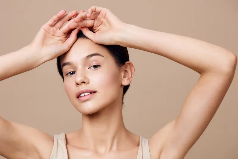 Young woman with natural makeup poses gracefully against a soft beige background, showcasing beauty and confidence through gentle expression and elegant posture
